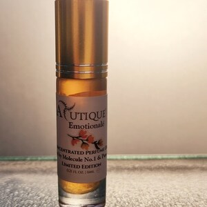 Puede incluir: Un frasco de perfume roll-on con tapa dorada y base de vidrio transparente. La etiqueta dice "AUTIQUE Emotionale" con texto debajo que indica que es un aceite de perfume concentrado. El frasco contiene 6 ml de perfume.