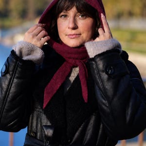 Könnte beinhalten: Ein dunkelburgunderfarbener Kapuzenschal, der am Hals gebunden ist und über einem beigen Strickpullover sowie einem schwarzen Ledermantel mit schwarzem Kunstfellkragen und Gürtel getragen wird. Der Schal hat einen strukturierten Strickstoff.