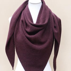 Könnte beinhalten: Ein dreieckiger Schal in tiefem Pflaumenton mit weicher Strickstruktur. Der Schal ist um eine weiße Schaufensterpuppe drapiert und zeichnet sich durch eine spitz zulaufende Unterkante sowie lange, schmal zulaufende Seiten aus, die herabhängen.