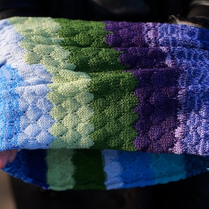 Op de afbeelding: Een gebreide armwarmer in tinten blauw, groen en paars. De stof heeft een textuur met een diamantachtig patroon. De armwarmer is opgevouwen, waardoor de binnenkant zichtbaar is. De achtergrond is donker, wat de kleuren van het gebreide item benadrukt.