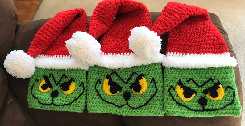 May include: Three crocheted Grinch hats with red and white Santa hats on top. The Grinch faces are green with yellow eyes and black eyebrows.