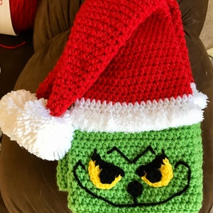 May include: Green crocheted Grinch face blanket with a red crocheted Santa hat on top. The hat has a white fur trim.