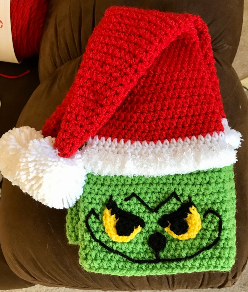 May include: Green crocheted Grinch face blanket with a red crocheted Santa hat on top. The hat has a white fur trim.