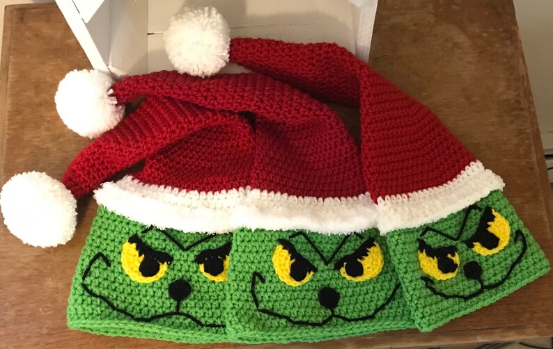 May include: Three crocheted Grinch hats with white pom-poms. The hats are red and green with a white trim and feature a Grinch face with yellow eyes and a black nose.