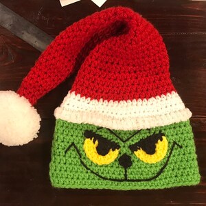 May include: Green and red crocheted Grinch hat with a white pom-pom on top. The hat features a Grinch face with yellow eyes and a black nose.
