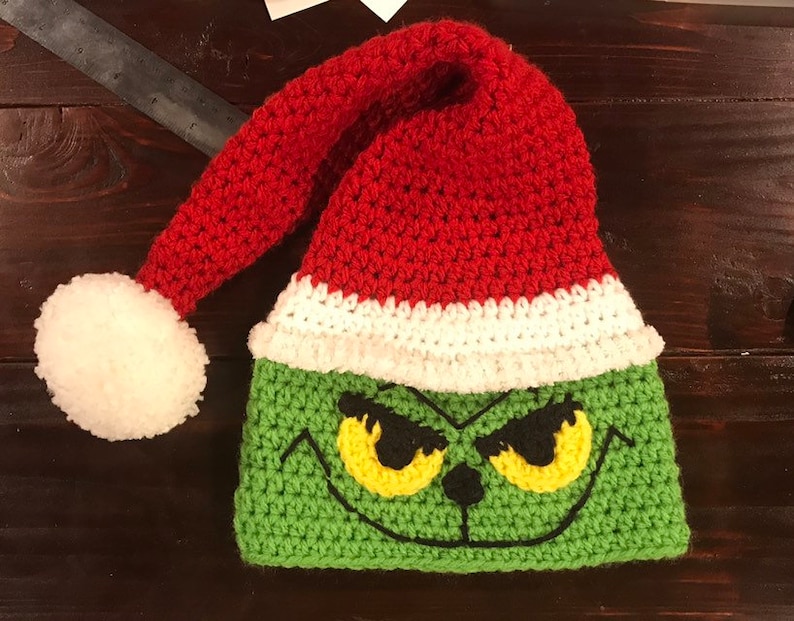 May include: Green and red crocheted Grinch hat with a white pom-pom on top. The hat features a Grinch face with yellow eyes and a black nose.