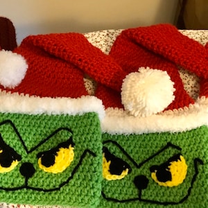 May include: Four crocheted Grinch-themed Christmas hats with red and white trim. Each hat features a green Grinch face with yellow eyes and a black nose.