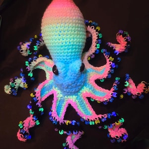 May include: A colorful, crocheted octopus with a gradient head in pink, blue, and green. The tentacles are black with pink and blue accents, adorned with glowing beads. The octopus has large, black button eyes.
