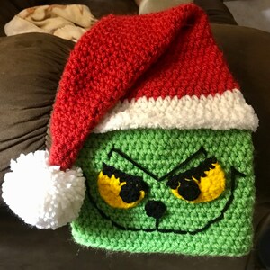 May include: Green crocheted Grinch hat with a red and white Santa hat on top. The Grinch has yellow eyes and a black nose.