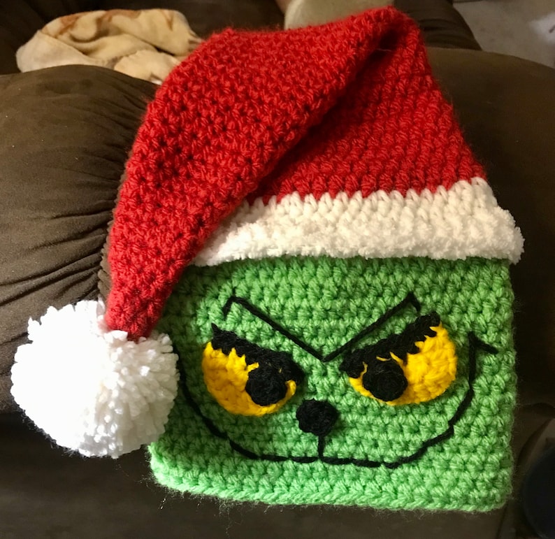 May include: Green crocheted Grinch hat with a red and white Santa hat on top. The Grinch has yellow eyes and a black nose.