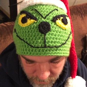 May include: Green crocheted Grinch hat with a red and white Santa hat on top. The hat has yellow eyes, a black nose, and a black stitched smile.