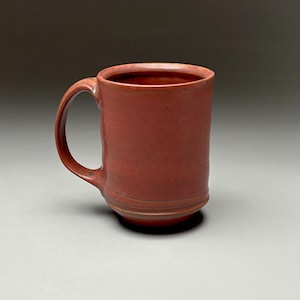 May include: A handmade, reddish-brown ceramic mug with a rounded handle. The mug has a cylindrical shape with a slightly flared rim and a decorative band near the base. The mug is suitable for hot or cold beverages.