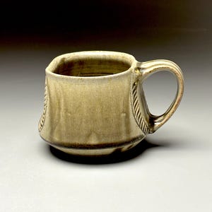 May include: A handmade ceramic mug with a square shape and a rounded handle. The mug has a textured, earthy appearance with a light brown glaze and decorative rope-like detailing on the side. The mug is approximately 4 inches tall.