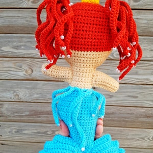 May include: A crocheted mermaid doll with red hair, a yellow crown, and a blue tail. The doll is holding a white pearl in each hand.