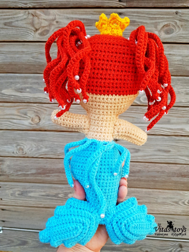 May include: A crocheted mermaid doll with red hair, a yellow crown, and a blue tail. The doll is holding a white pearl in each hand.