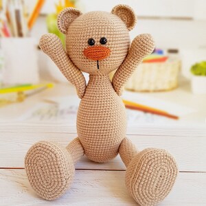 May include: A crocheted beige teddy bear with black eyes and an orange nose, sitting with its arms raised and legs outstretched on a white wooden surface.