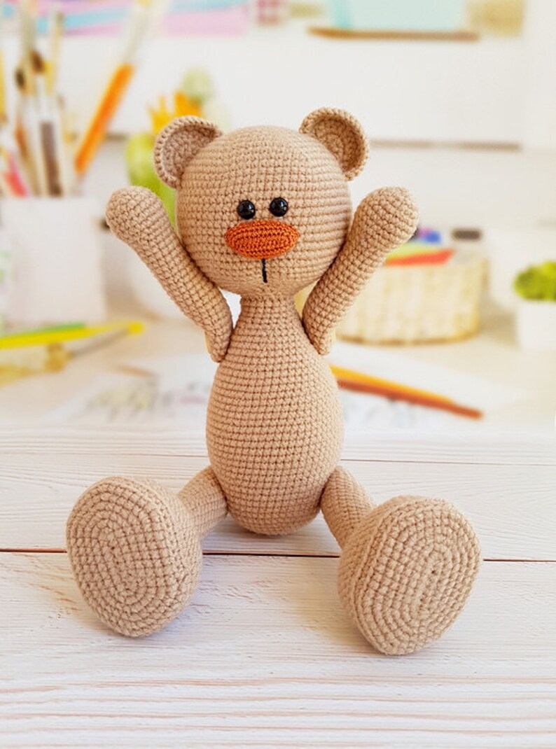 May include: A crocheted beige teddy bear with black eyes and an orange nose, sitting with its arms raised and legs outstretched on a white wooden surface.
