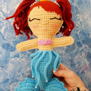 May include: A crocheted mermaid doll with red hair, a yellow crown, and a blue tail. The doll is wearing a pink seashell top and has a sweet expression on its face.