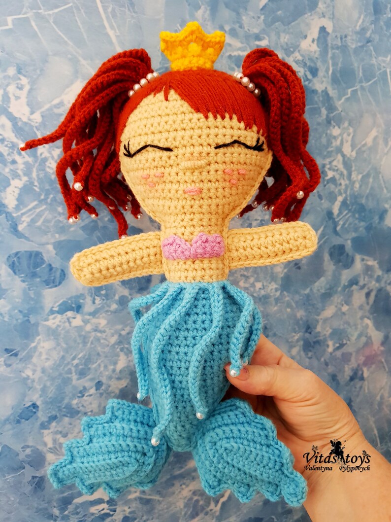 May include: A crocheted mermaid doll with red hair, a yellow crown, and a blue tail. The doll is wearing a pink seashell top and has a sweet expression on its face.
