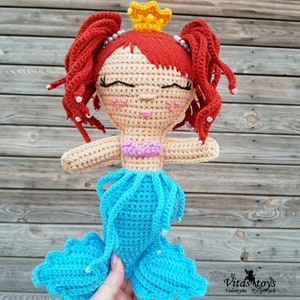 May include: A crocheted mermaid doll with red hair, a yellow crown, and a blue tail. The doll has a pink top and white beads around her head. The doll is holding a hand with a white bead on her finger.