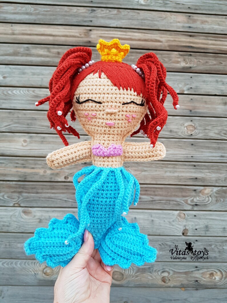 May include: A crocheted mermaid doll with red hair, a yellow crown, and a blue tail. The doll has a pink top and white beads around her head. The doll is holding a hand with a white bead on her finger.