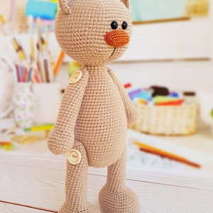 May include: A beige crocheted teddy bear with a brown nose and black eyes. The bear has wooden buttons for joints.
