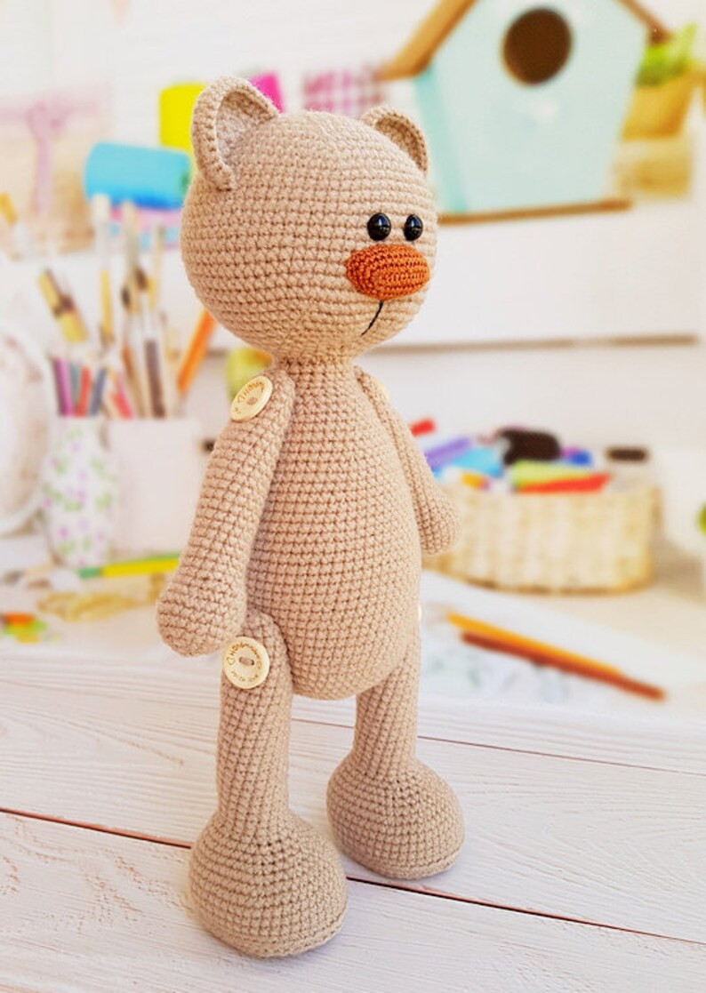 May include: A beige crocheted teddy bear with a brown nose and black eyes. The bear has wooden buttons for joints.