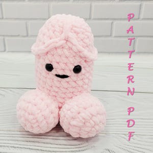 May include: A plush, pink, phallic-shaped toy with a smiling face and two round testicles. The toy is made of a soft, textured material. The word "PATTERN PDF" is written vertically in pink letters.