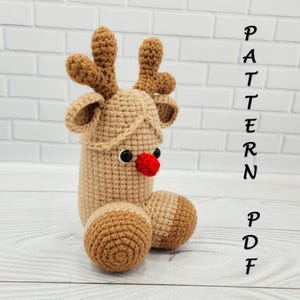 May include: A handmade crochet reindeer figure in shades of brown, with a red nose and black eyes. The reindeer has large antlers and a rounded body. The word "PATTERN PDF" is written vertically in black.