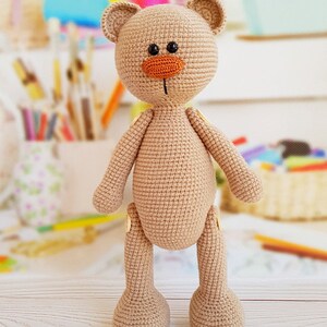 May include: A crocheted brown teddy bear with a large orange nose and black eyes. The bear is standing on a light wood surface.