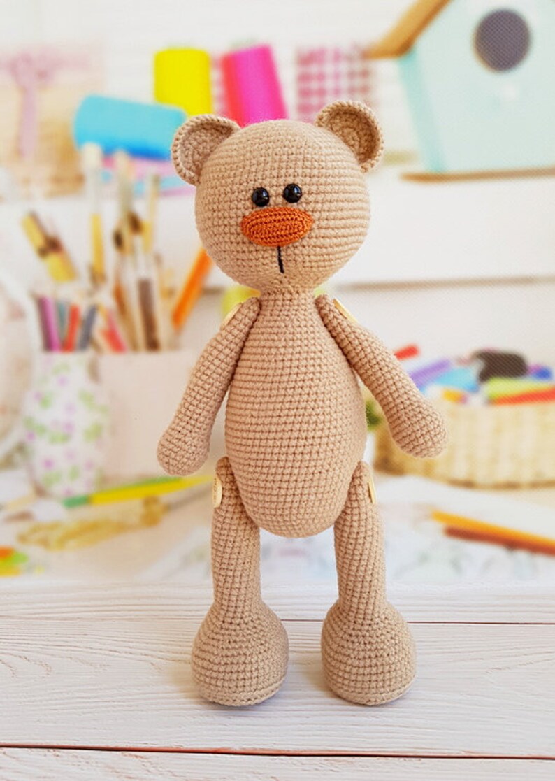 May include: A crocheted brown teddy bear with a large orange nose and black eyes. The bear is standing on a light wood surface.