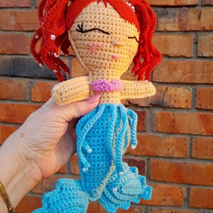 May include: Crocheted mermaid doll with red hair, a yellow crown, and a blue tail. The doll has a pink top and white pearl accents.