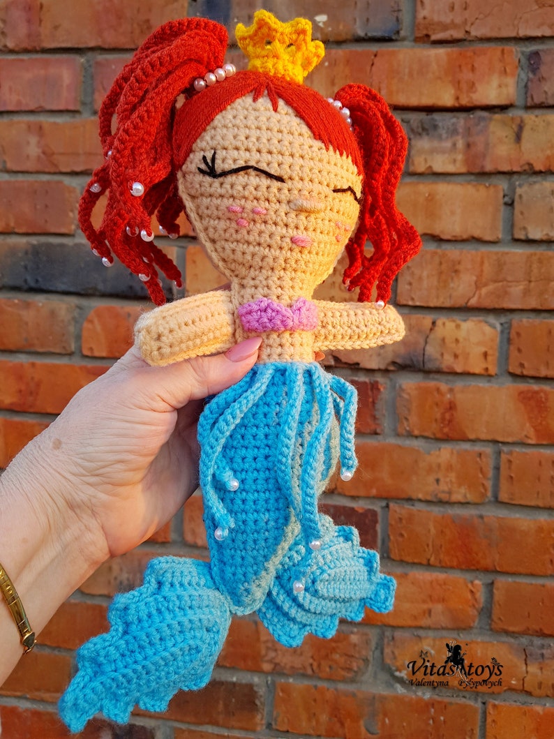 May include: Crocheted mermaid doll with red hair, a yellow crown, and a blue tail. The doll has a pink top and white pearl accents.