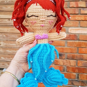 May include: A crocheted mermaid doll with red hair, a yellow crown, and a blue tail. The doll has a pink top and white beads around her hair and tail.
