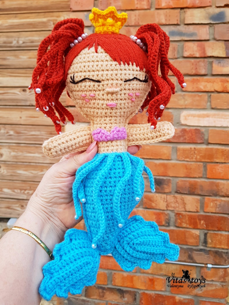 May include: A crocheted mermaid doll with red hair, a yellow crown, and a blue tail. The doll has a pink top and white beads around her hair and tail.