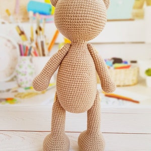 May include: A crocheted beige teddy bear standing on a white wooden surface. The bear has a simple design with no facial features.