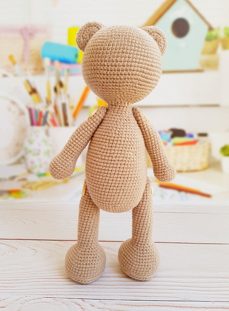 May include: A crocheted beige teddy bear standing on a white wooden surface. The bear has a simple design with no facial features.