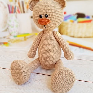May include: A crocheted beige teddy bear with orange nose and black eyes sitting on a white wooden surface.