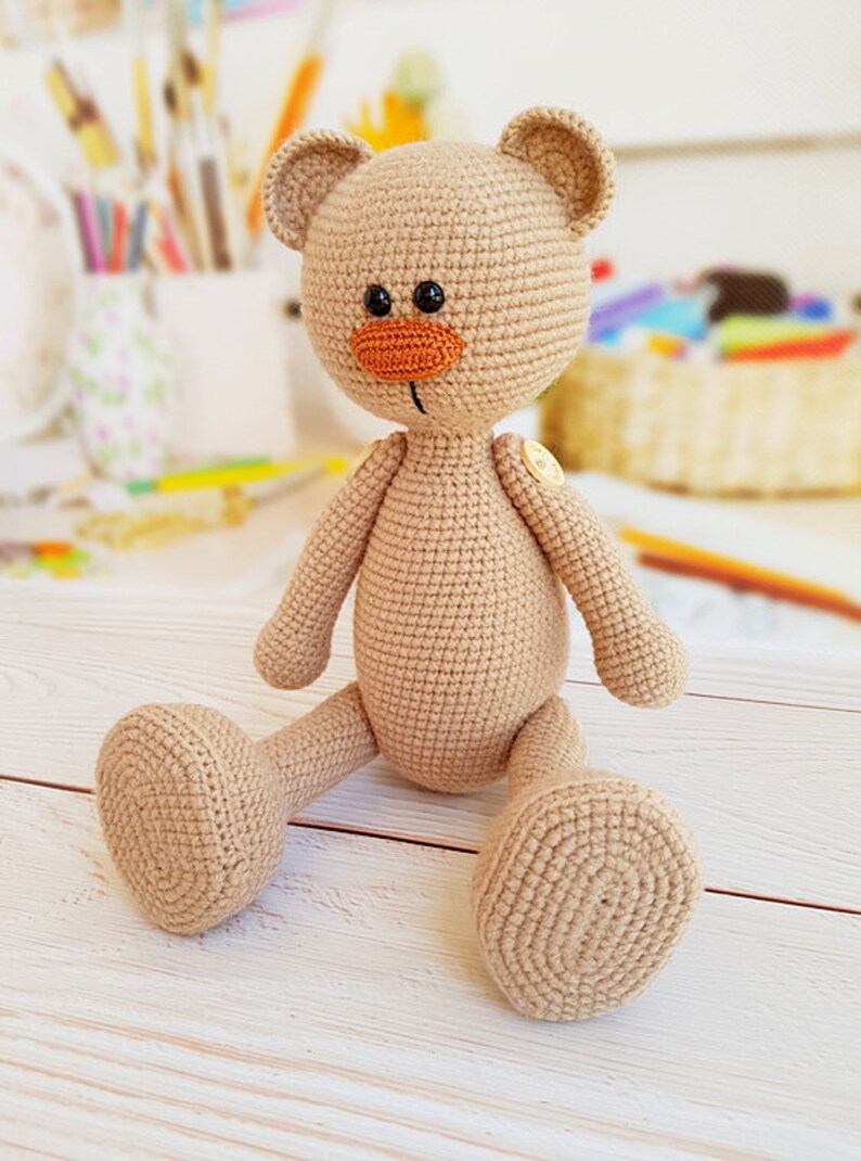 May include: A crocheted beige teddy bear with orange nose and black eyes sitting on a white wooden surface.
