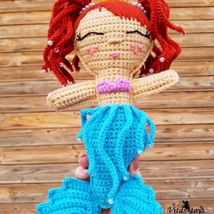 May include: A crocheted mermaid doll with red hair, a yellow crown, and a blue tail. The doll is wearing a pink bikini top and has white pearl accents.