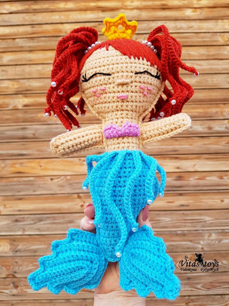 May include: A crocheted mermaid doll with red hair, a yellow crown, and a blue tail. The doll is wearing a pink bikini top and has white pearl accents.