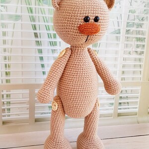 May include: A crocheted beige teddy bear with button joints. The bear has a brown nose and black eyes.