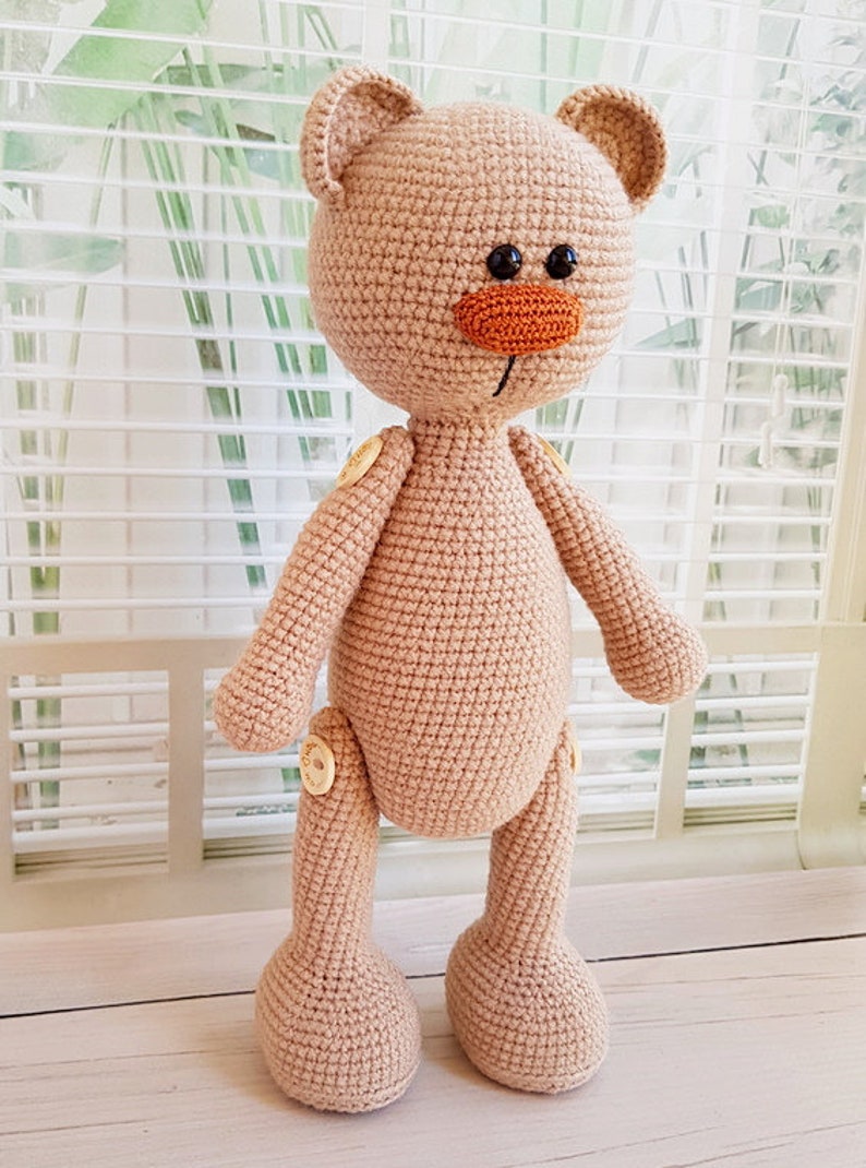May include: A crocheted beige teddy bear with button joints. The bear has a brown nose and black eyes.