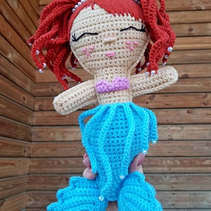 May include: A crocheted mermaid doll with a yellow crown, red hair, and a blue tail. The doll has a pink and purple top and is holding a pearl in its hand.