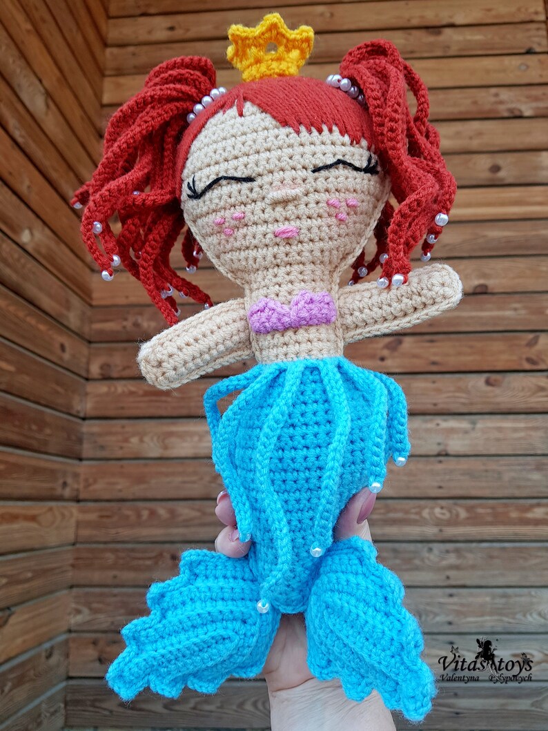 May include: A crocheted mermaid doll with a yellow crown, red hair, and a blue tail. The doll has a pink and purple top and is holding a pearl in its hand.