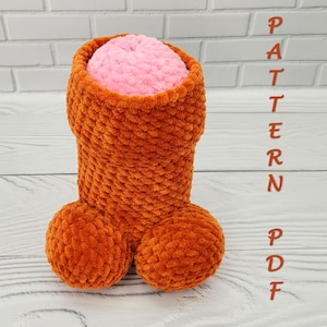 May include: A handmade, plush, orange-brown penis-shaped item with a pink interior. The item has two round balls at the base and a cylindrical shaft. The word "PATTERN PDF" is written vertically in orange.