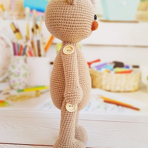 May include: A beige crocheted teddy bear with brown button eyes and a brown nose. The bear has two brown buttons on its body and is standing on a white wooden surface.