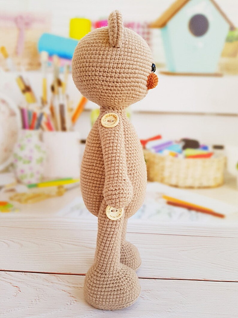 May include: A beige crocheted teddy bear with brown button eyes and a brown nose. The bear has two brown buttons on its body and is standing on a white wooden surface.