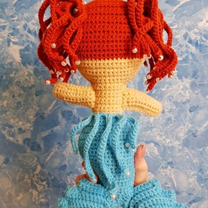 May include: A crocheted mermaid doll with red hair, a yellow crown, and a blue tail. The doll is wearing a white pearl necklace and has a light blue and yellow bodice.