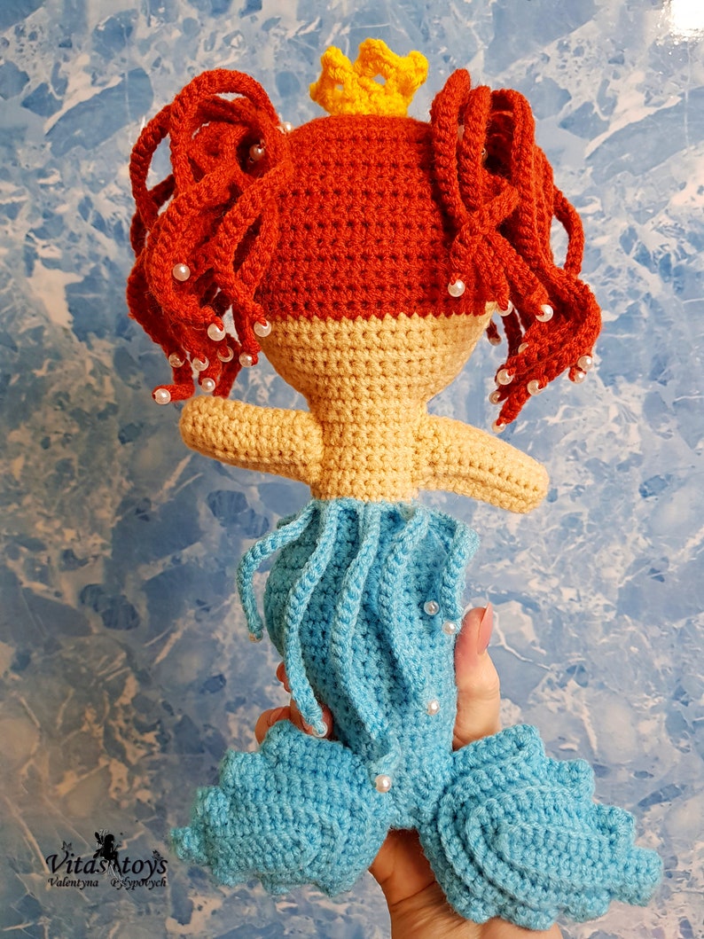 May include: A crocheted mermaid doll with red hair, a yellow crown, and a blue tail. The doll is wearing a white pearl necklace and has a light blue and yellow bodice.